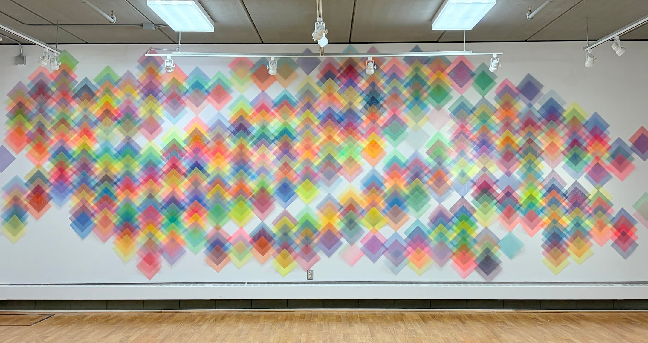 Colorful abstract wall installation composed of hundreds of overlapping translucent diamond shapes arranged in a flowing pattern across a large white gallery wall.