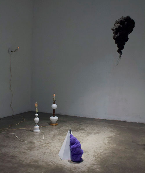 Minimalist installation in a gallery space featuring sculptural objects on the floor, including lamps, cords, and purple textured forms, with a dark suspended shape on the wall above.