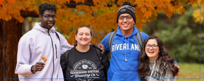 Four smiling Riverland students stand together outdoors in front of vibrant autumn leaves, dressed in casual fall clothing.
