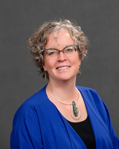 Studio shot of Kat Linaker, Riverland President, smiling, wearing glasses with a blue top and pendant necklace